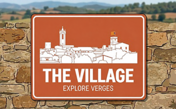 White outline of Verges on a terracotta sign in front of a stone wall, reading “The Village · Explore Verges”.