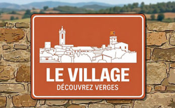 Silhouette de Verges sur un panneau devant un mur de pierre, avec le texte «Le village · Découvrez Verges».