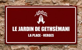 Mains blanches jointes sous une branche d'olivier sur un panneau rouge «Le Jardin de Gethsémani · La Place, Verges».