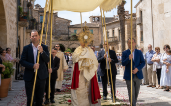Procesión de Corpus Christi con custodia, palio y participantes en una calle empedrada.