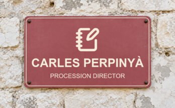 Dusty-rose sign on a limestone wall reading “CARLES PERPINYÀ”, with a pictogram.