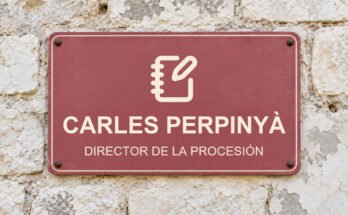 Rótulo rosa sobre un muro de piedra caliza con el texto «CARLES PERPINYÀ» y un pictograma.