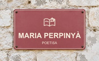 Rótulo rosa sobre un muro de piedra caliza con el texto «MARIA PERPINYÀ» y un pictograma.