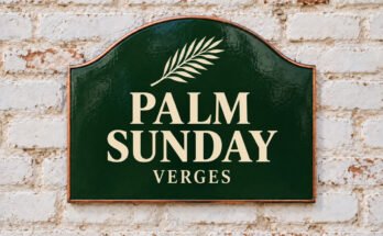 Arched green sign reading “PALM SUNDAY”, with a palm branch, on a whitewashed brick wall.