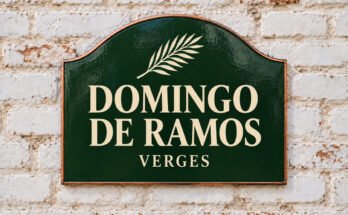 Rótulo verde arqueado con «DOMINGO DE RAMOS» y una rama de palma, sobre un muro de ladrillo encalado.