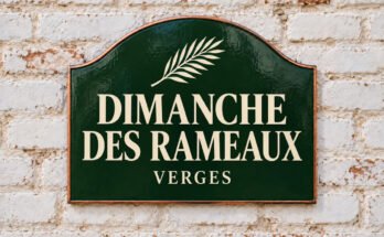 Plaque verte cintrée portant « DIMANCHE DES RAMEAUX » et une branche de palmier, sur un mur de briques blanchies.