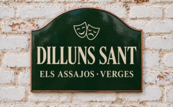Rètol verd arquejat amb «DILLUNS SANT» i màscares de teatre, sobre un mur de maó emblanquinat.