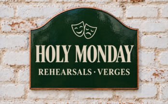 Arched green sign reading “HOLY MONDAY”, with theatre masks, on a whitewashed brick wall.