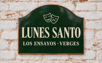 Rótulo verde arqueado con «LUNES SANTO» y máscaras de teatro, sobre un muro de ladrillo encalado.