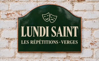 Plaque verte cintrée portant « LUNDI SAINT » et des masques de théâtre, sur un mur de briques blanchies.