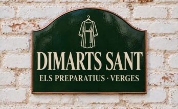 Rètol verd arquejat amb «DIMARTS SANT» i un vestit penjat, sobre un mur de maó emblanquinat.