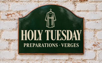 Arched green sign reading “HOLY TUESDAY”, with a hanging tunic, on a whitewashed brick wall.