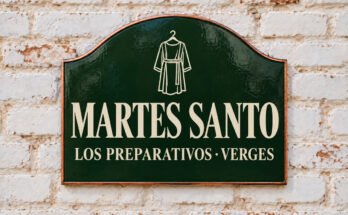 Rótulo verde arqueado con «MARTES SANTO» y una túnica colgada, sobre un muro de ladrillo encalado.