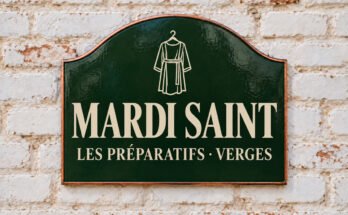 Plaque verte cintrée portant « MARDI SAINT » et une tunique suspendue, sur un mur de briques blanchies.