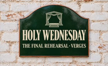 Arched green sign reading “HOLY WEDNESDAY”, with a stage with curtains, on a whitewashed brick wall.