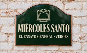 Rótulo verde arqueado con «MIÉRCOLES SANTO» y un escenario con cortinas, sobre un muro de ladrillo encalado.