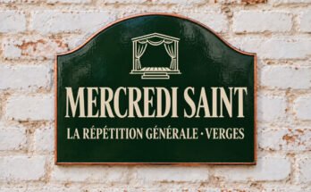 Plaque verte cintrée portant « MERCREDI SAINT » et une scène avec des rideaux, sur un mur de briques blanchies.