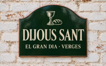 Rètol verd arquejat amb «DIJOUS SANT» i pa i un calze, sobre un mur de maó emblanquinat.