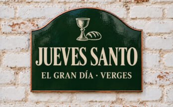 Rótulo verde arqueado con «JUEVES SANTO» y pan y un cáliz, sobre un muro de ladrillo encalado.