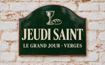 Plaque verte cintrée portant « JEUDI SAINT » et du pain et un calice, sur un mur de briques blanchies.