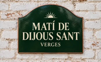 Rètol verd arquejat amb «MATÍ DE DIJOUS SANT» i un sol naixent, sobre un mur de maó emblanquinat.