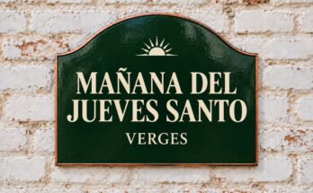 Rótulo verde arqueado con «MAÑANA DEL JUEVES SANTO» y un sol naciente, sobre un muro de ladrillo encalado.