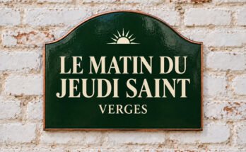 Plaque verte cintrée portant « LE MATIN DU JEUDI SAINT » et un soleil levant, sur un mur de briques blanchies.