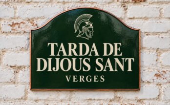 Rètol verd arquejat amb «TARDA DE DIJOUS SANT» i un casc romà, sobre un mur de maó emblanquinat.