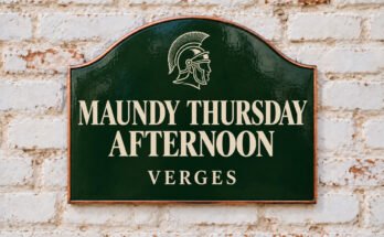 Arched green sign reading “MAUNDY THURSDAY AFTERNOON”, with a Roman helmet, on a whitewashed brick wall.