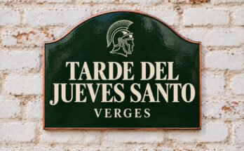 Rótulo verde arqueado con «TARDE DEL JUEVES SANTO» y un casco romano, sobre un muro de ladrillo encalado.