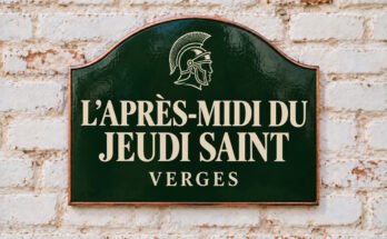Plaque verte cintrée portant « L’APRÈS-MIDI DU JEUDI SAINT » et un casque romain, sur un mur de briques blanchies.