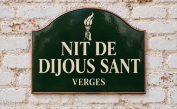 Rètol verd arquejat amb «NIT DE DIJOUS SANT» i una torxa encesa, sobre un mur de maó emblanquinat.