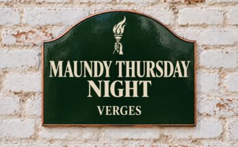 Arched green sign reading “MAUNDY THURSDAY NIGHT”, with a lit torch, on a whitewashed brick wall.