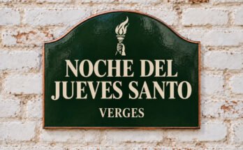Rótulo verde arqueado con «NOCHE DEL JUEVES SANTO» y una antorcha encendida, sobre un muro de ladrillo encalado.