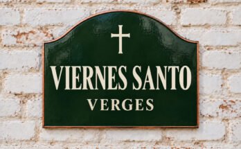 Rótulo verde arqueado con «VIERNES SANTO» y una cruz, sobre un muro de ladrillo encalado.