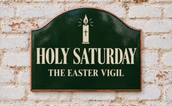 Arched green sign reading “HOLY SATURDAY”, with an Easter candle, on a whitewashed brick wall.