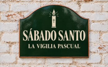 Rótulo verde arqueado con «SÁBADO SANTO» y un cirio pascual, sobre un muro de ladrillo encalado.