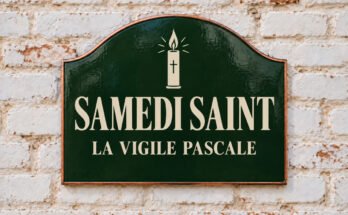 Plaque verte cintrée portant « SAMEDI SAINT » et un cierge pascal, sur un mur de briques blanchies.