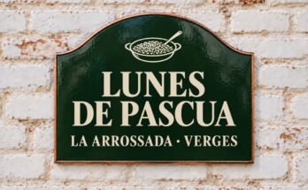 Rótulo verde arqueado con «LUNES DE PASCUA» y un plato de arroz y una cuchara, sobre un muro de ladrillo encalado.