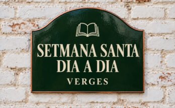 Rètol verd arquejat amb «SETMANA SANTA DIA A DIA» i un llibre obert, sobre un mur de maó emblanquinat.
