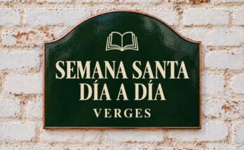 Rótulo verde arqueado con «SEMANA SANTA DÍA A DÍA» y un libro abierto, sobre un muro de ladrillo encalado.