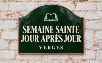 Plaque verte cintrée portant « SEMAINE SAINTE JOUR APRÈS JOUR » et un livre ouvert, sur un mur de briques blanchies.